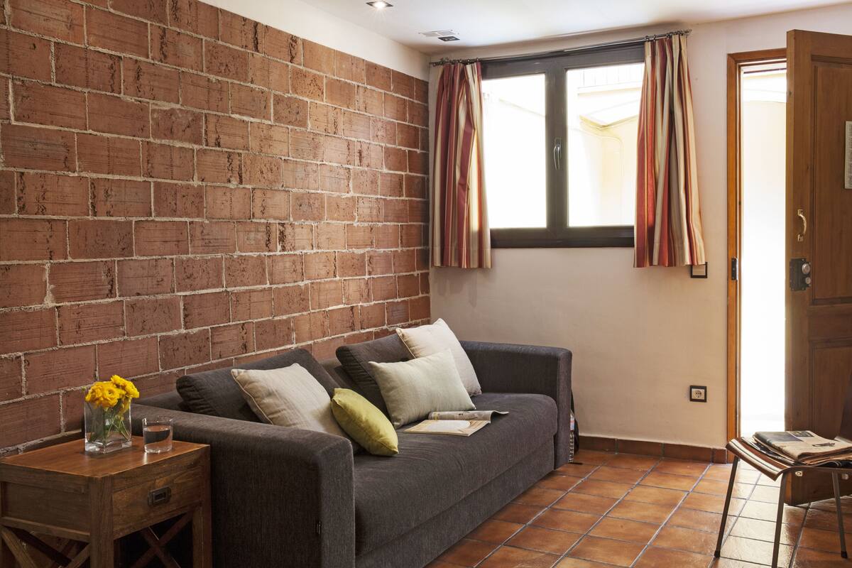 Een kamer met een bakstenen muur, een grijze bank met kussens, een houten bijzettafeltje met een bloemenvaas, en een raam met gordijnen.
