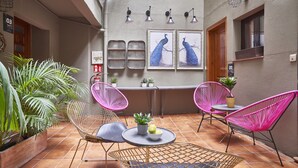 En patio med terracotta-fliser, to runde borde og fire pink metalstole. Der er plantekasser og to billeder i rammer på væggen.