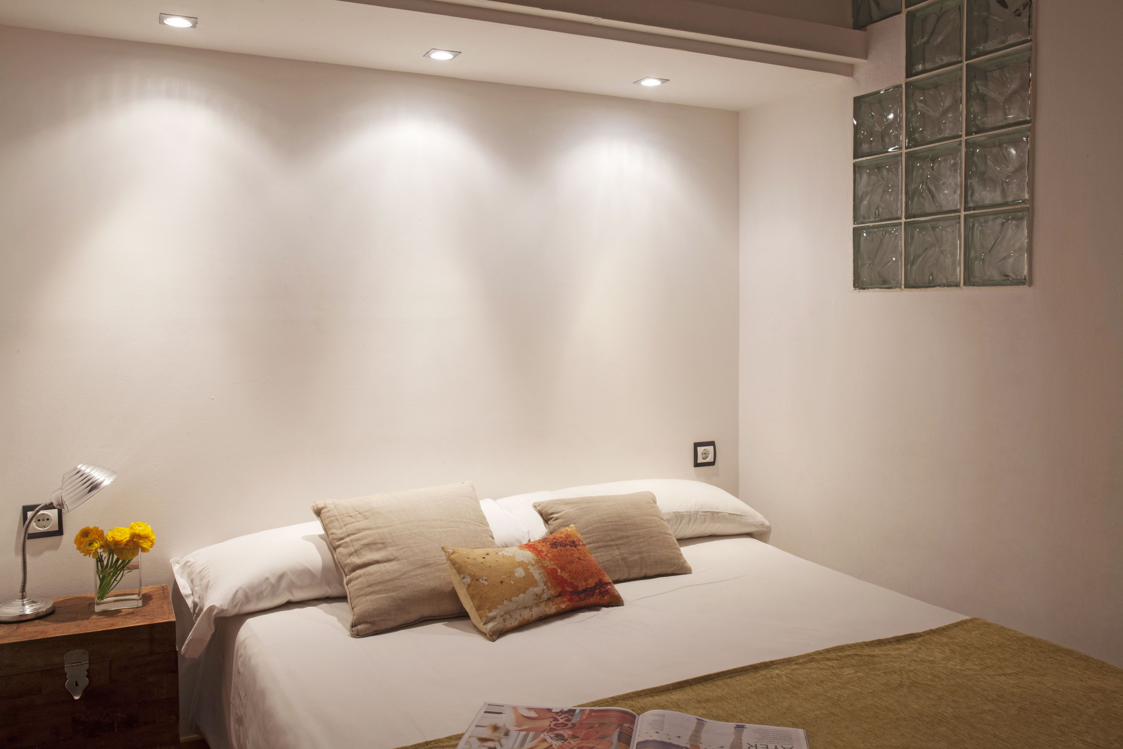 Habitación de hotel impecable con una cama, almohadas, una mesita de noche con lámpara y una revista sobre la cama.