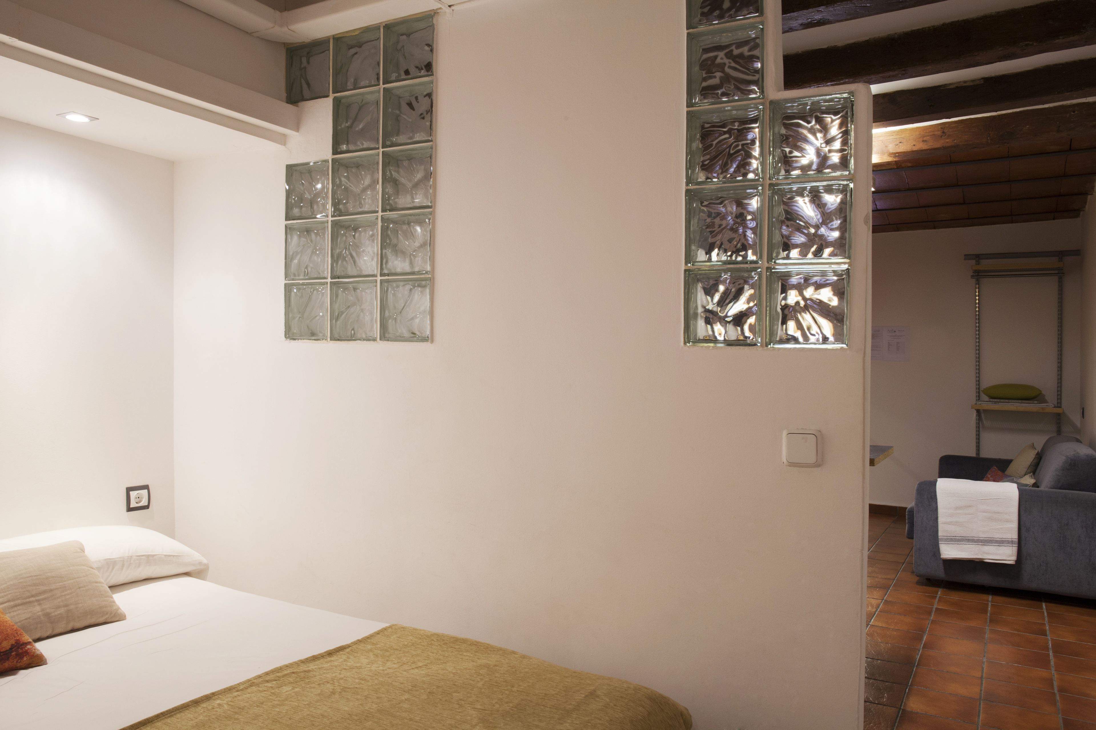 A bedroom with a bed, a window with decorative glass, and a view into a living area with a sofa.