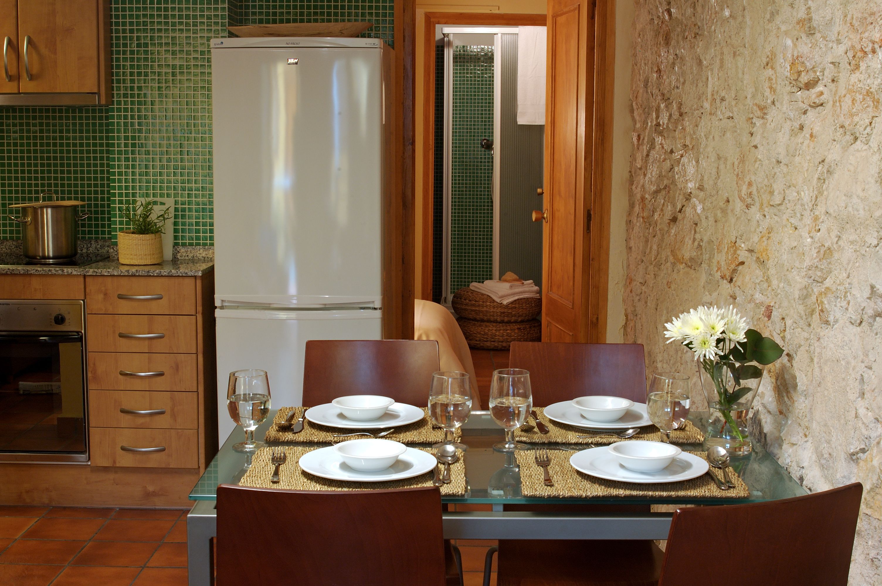 Een eethoek met een glazen tafel gedekt voor vier personen, omringd door houten stoelen. In de achtergrond is een koelkast en een fornuis zichtbaar.