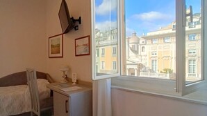 A hotel room with a bed, a desk, a chair, and a large window offering a city view. - Hotel Helvetia (Genoa)