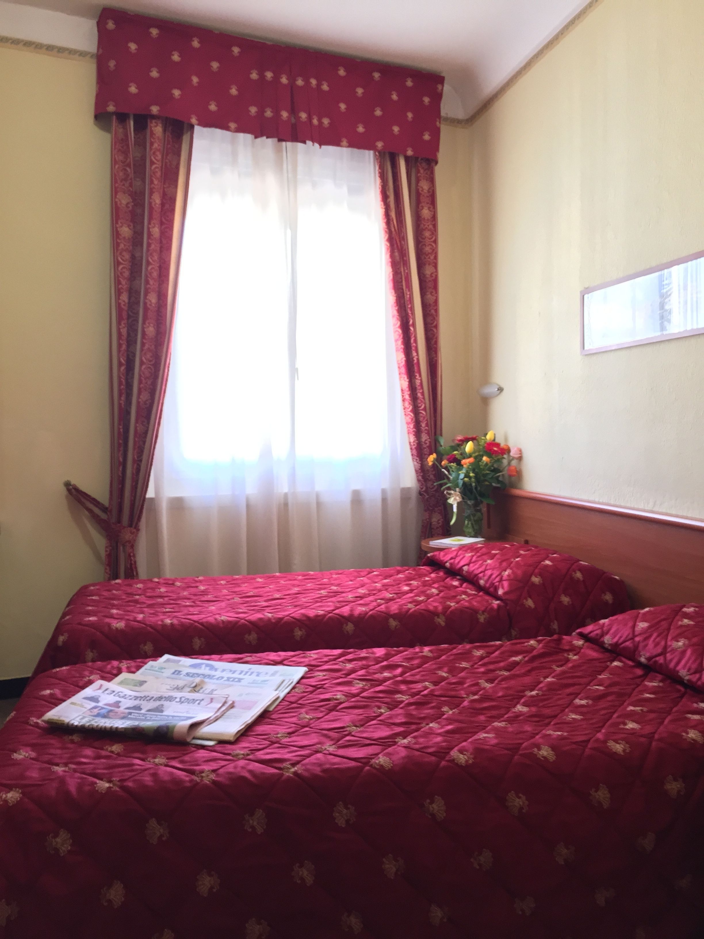 A hotel room with two beds, a window with curtains, a bedside table with a vase of flowers, and a framed picture on the wall.