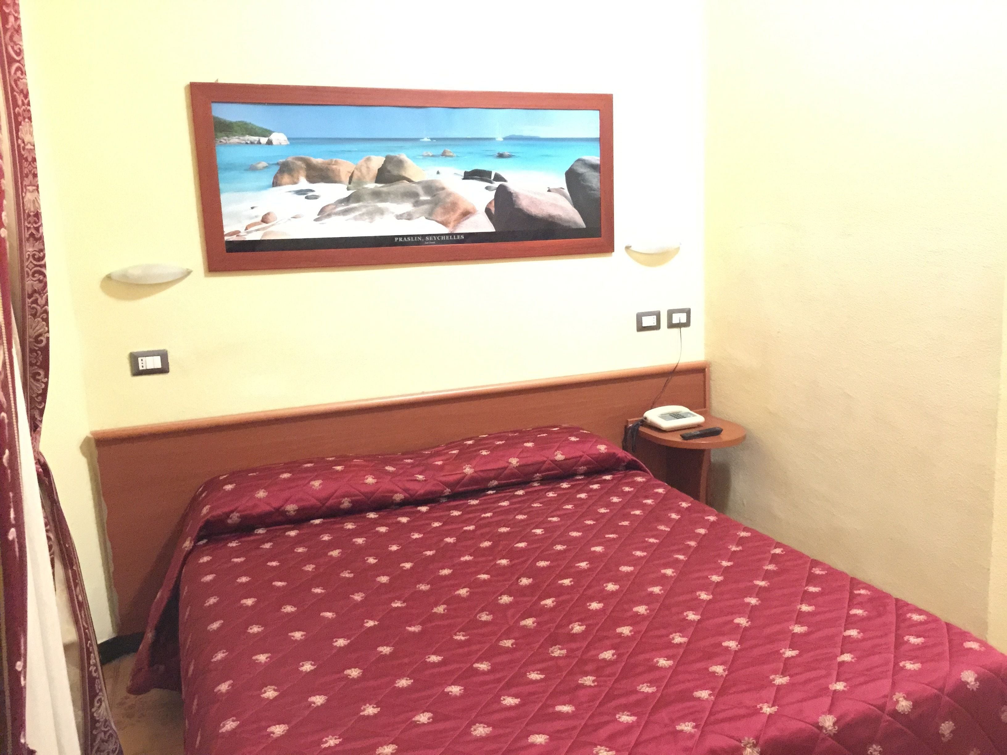 A hotel room with a bed, a wall-mounted TV displaying a beach scene, a bedside table with a phone and remote control, and a power outlet.
