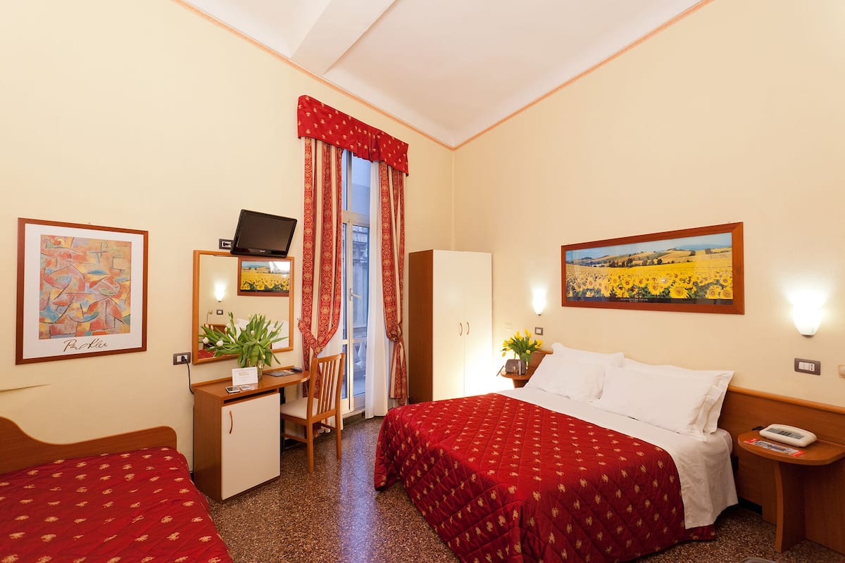 Habitación de hotel con dos camas, un escritorio, una silla, un televisor y obras de arte en las paredes.