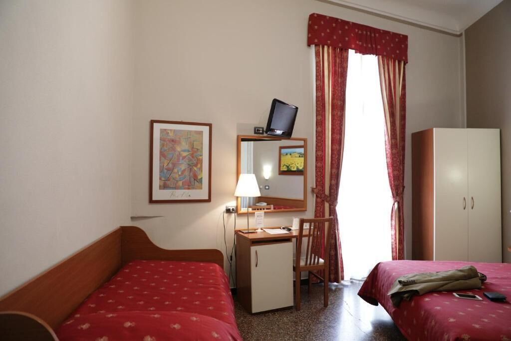 A hotel room with two single beds, a desk with a computer, a chair, a wardrobe, and a window with curtains.