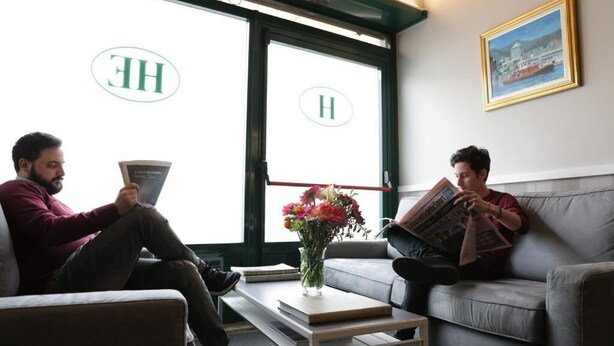 Two individuals seated on a grey sofa in a room with large windows, reading books. A vase with flowers is on a small table in front of them. - Hotel Helvetia (Genoa)