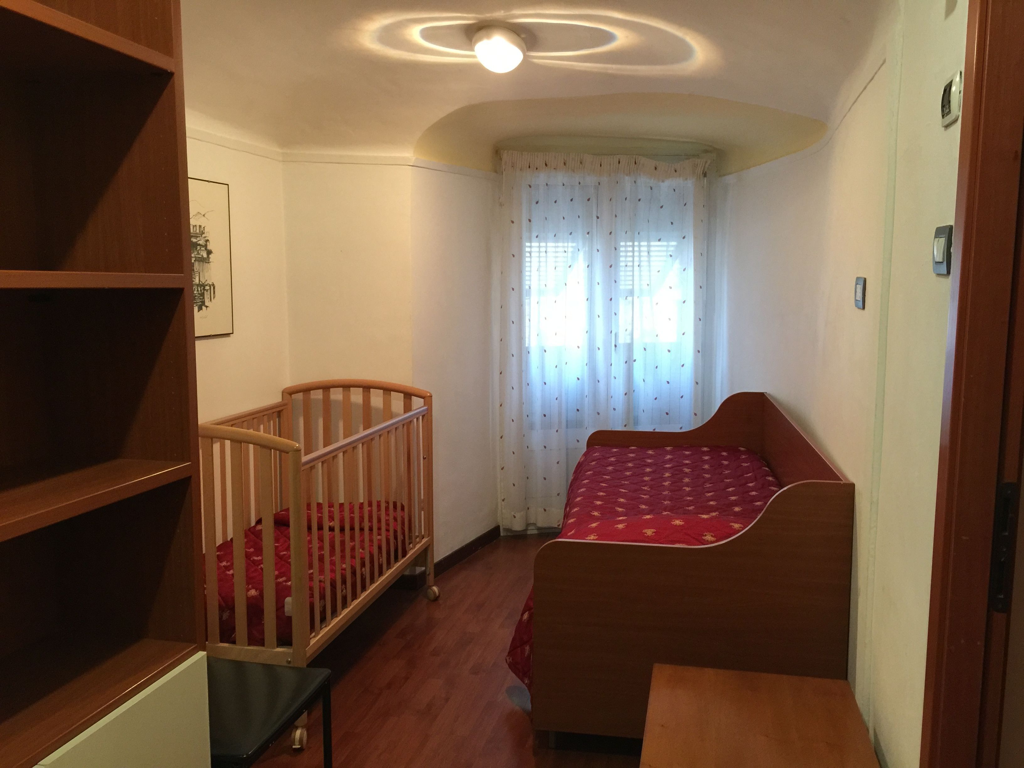 A room with a wooden crib, a small bed with a red bedspread, a wooden shelf unit, and a window with sheer curtains.