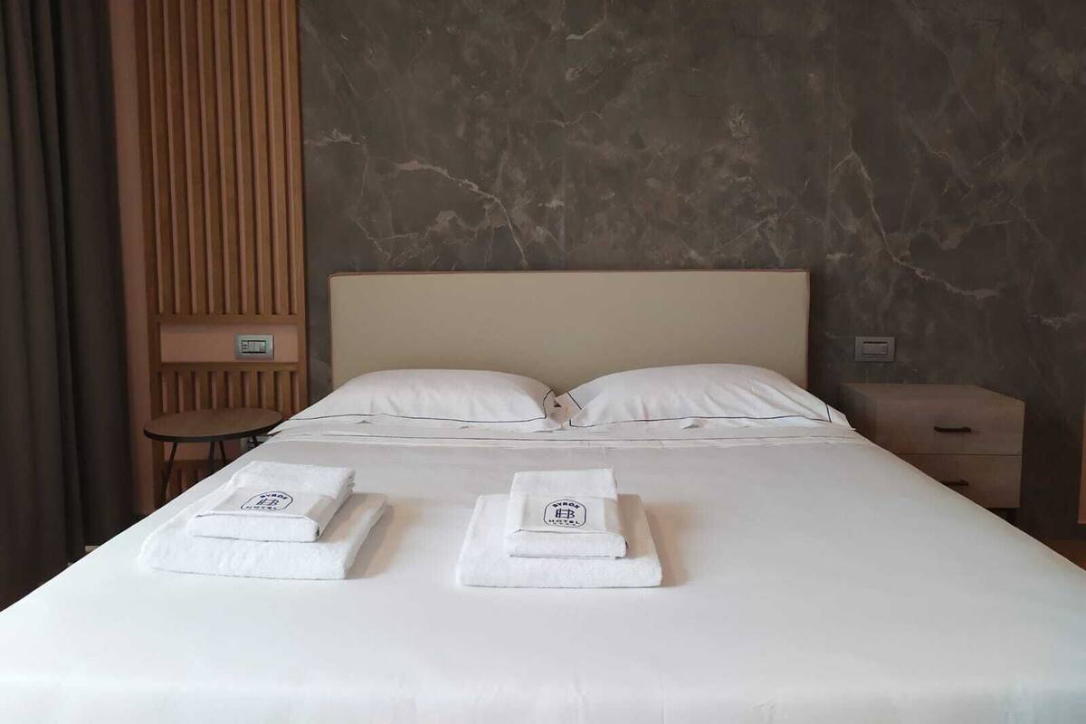 Une chambre d’hôtel avec un grand lit, deux oreillers, une table de chevet en bois, une petite table et un mur dont la texture ressemble à du marbre.