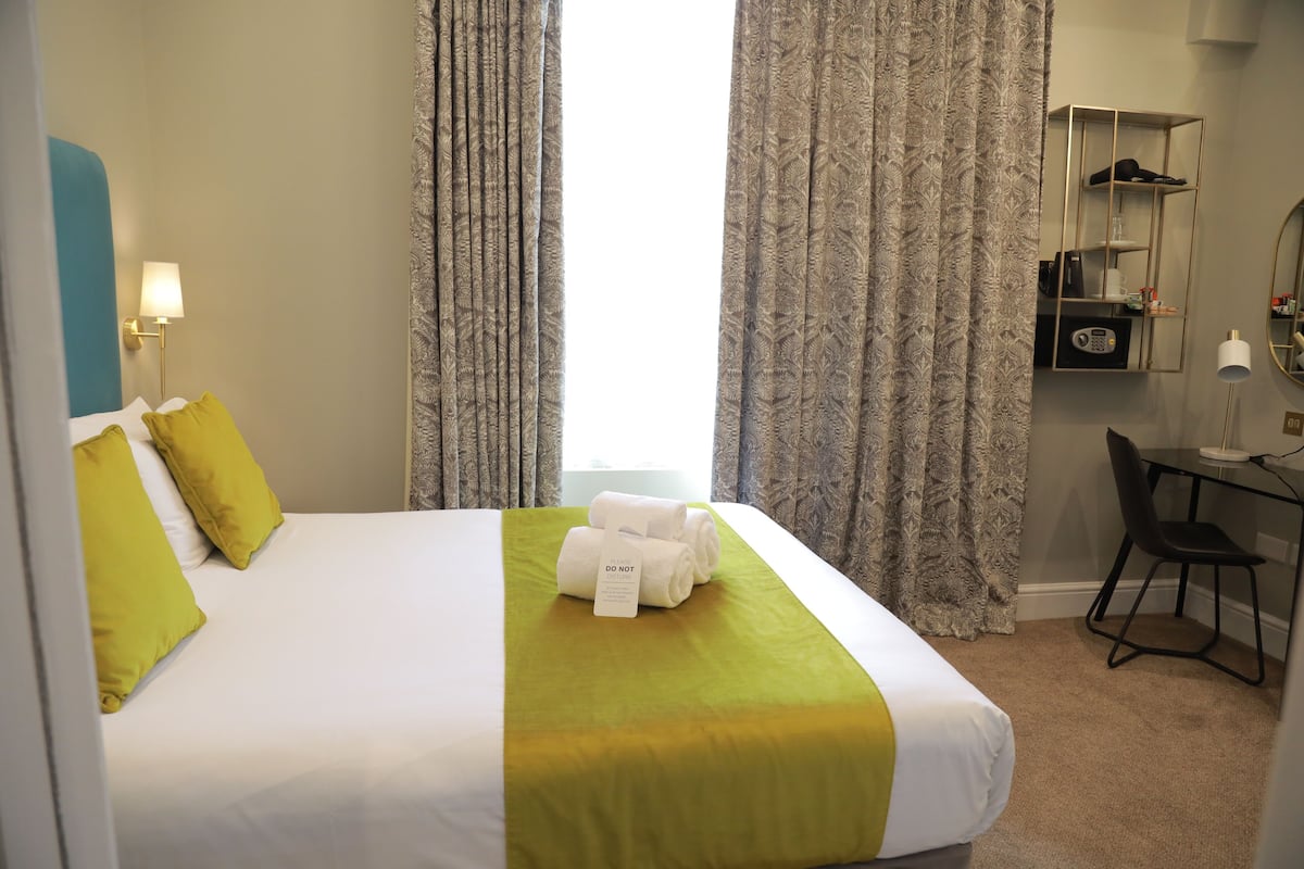 A hotel room with a neatly made bed, a desk with a chair, a mirror, and a shelf with personal items.