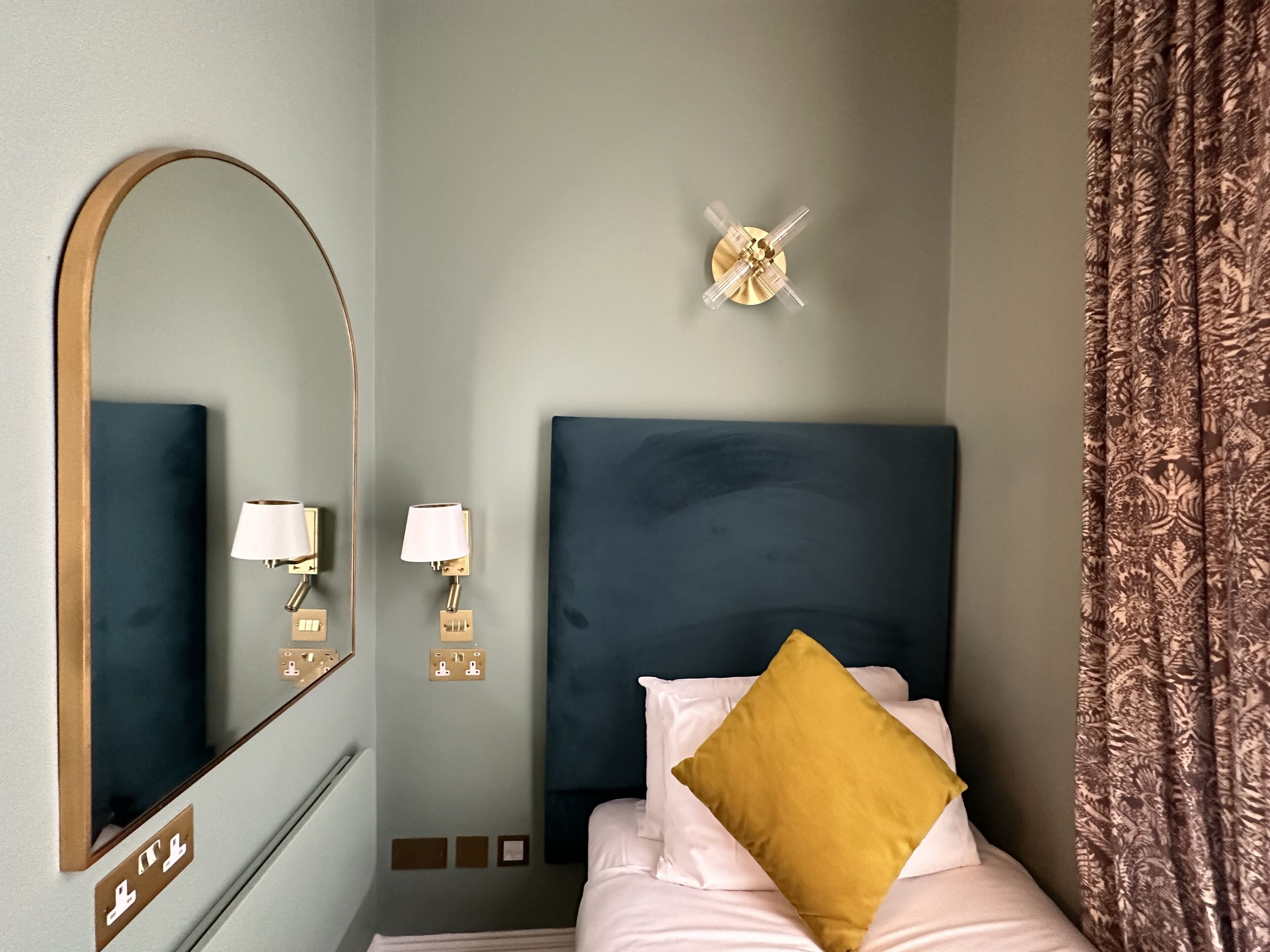 A hotel room with a large mirror, two wall-mounted lamps, a bed with white bedding and a mustard-colored pillow, and a patterned curtain.