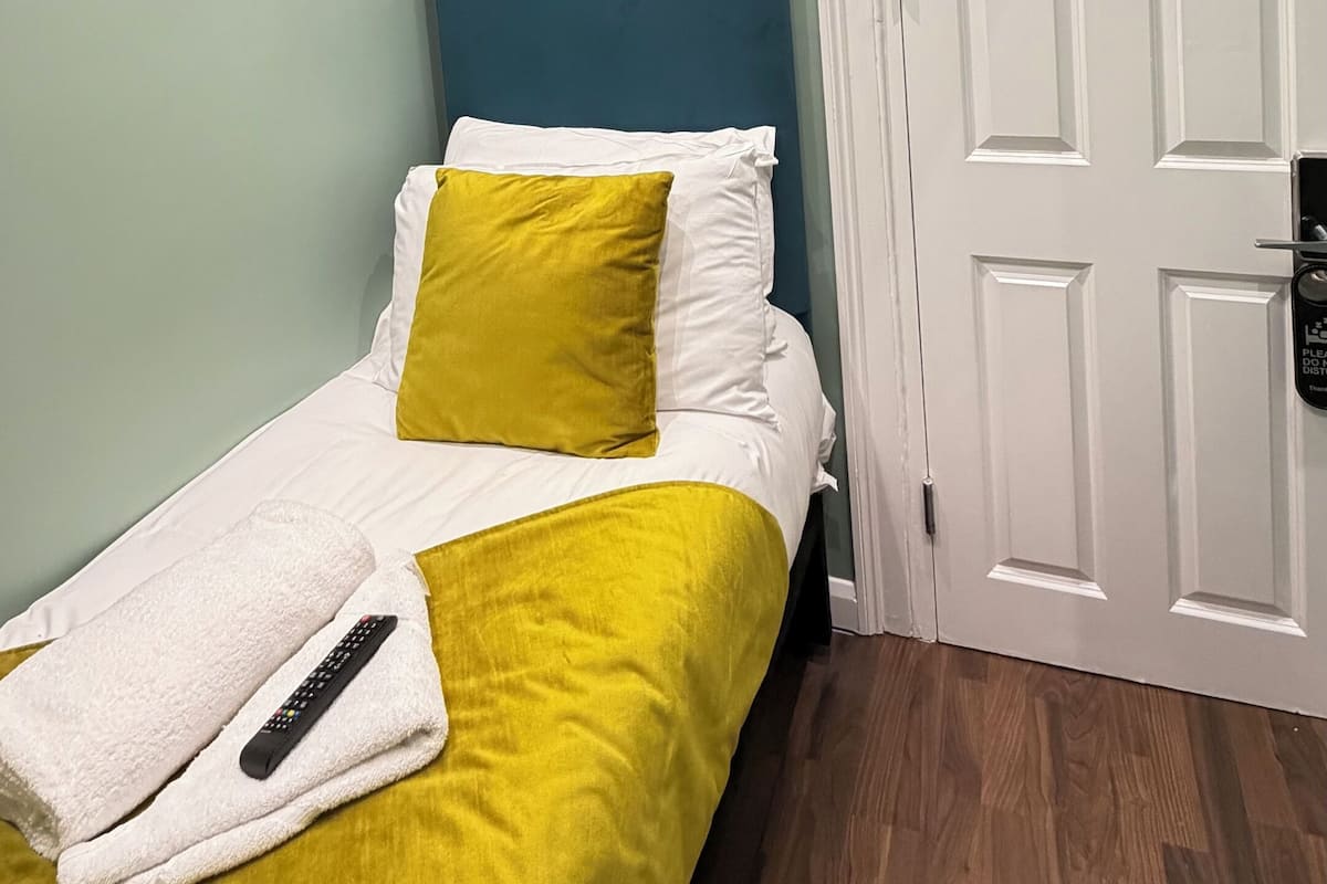 A bedroom with a bed, a remote control, a towel, a door with a lock, and a wall-mounted light fixture.