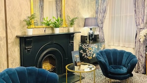 A living room with a fireplace, two blue armchairs, a round coffee table, and a chandelier. - Bloomsbury Palace Hotel (London)