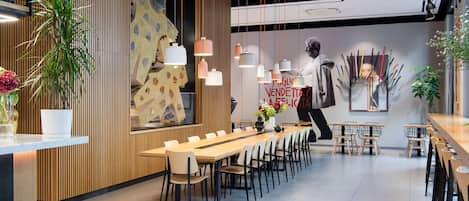 A modern restaurant interior with long communal tables, hanging pendant lights, and a large mural on the wall.