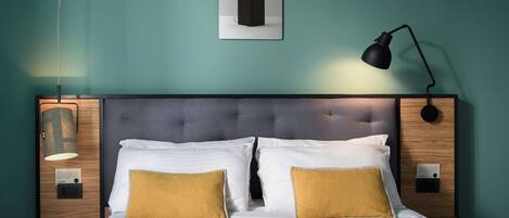 A neatly made bed with white and mustard pillows, a dark headboard, and a wall-mounted lamp.