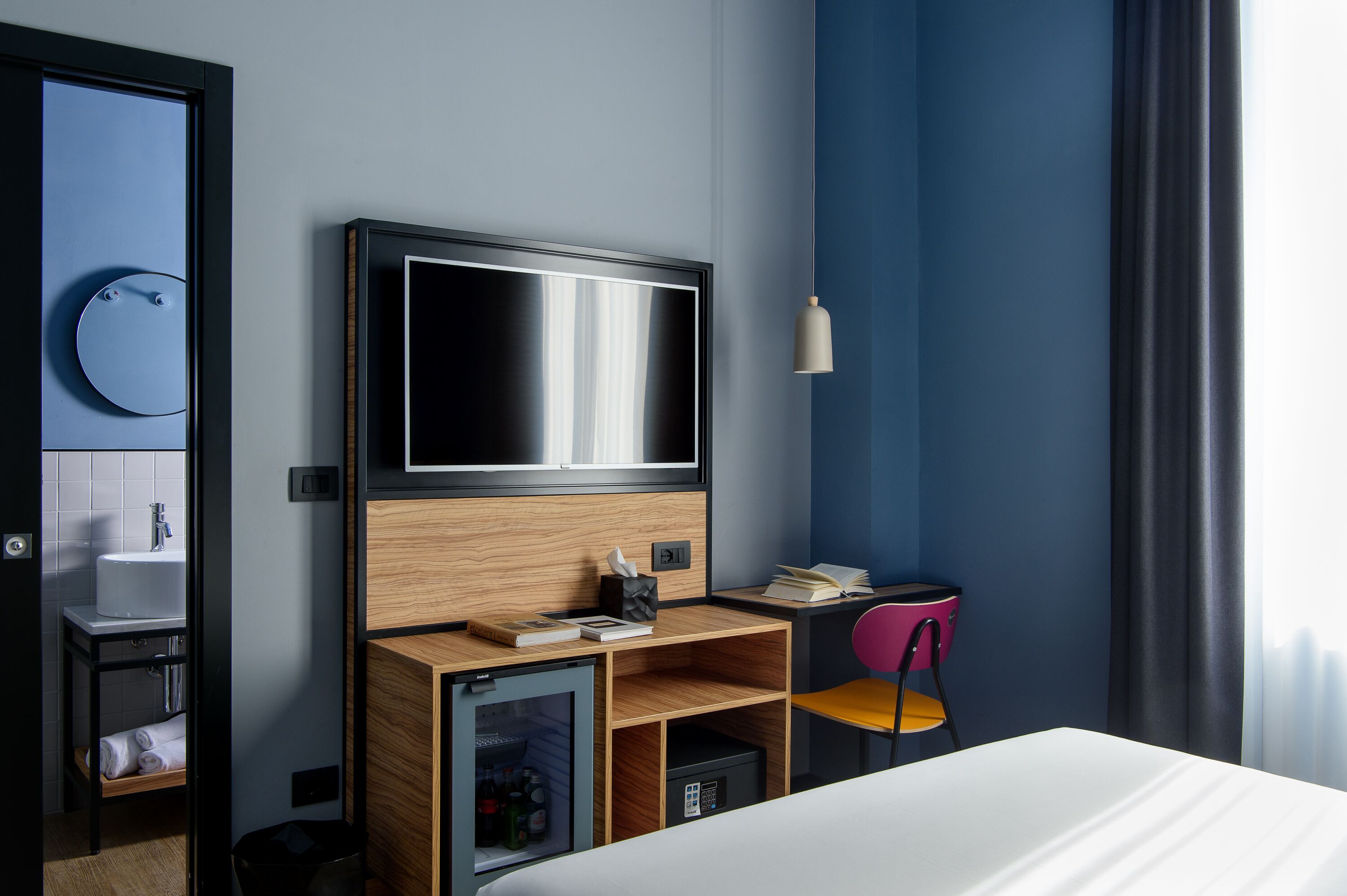 Una habitación de hotel moderna con un televisor de pantalla plana grande, un escritorio de madera, un refrigerador pequeño, una silla rosa y una mesita de noche con lámpara.