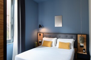 Una cama bien hecha con almohadas blancas y amarillas, una lámpara de noche y una mesita de madera con un libro y un frasco.