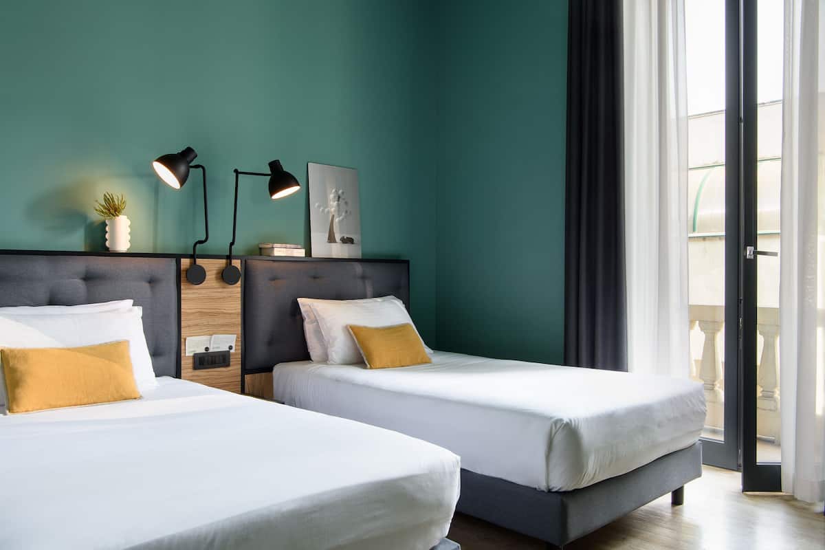 Een hotelkamer met twee bedden, een donker hoofdeinde, een kamerplant, een bureau met een lamp en uitzicht op de omgeving.