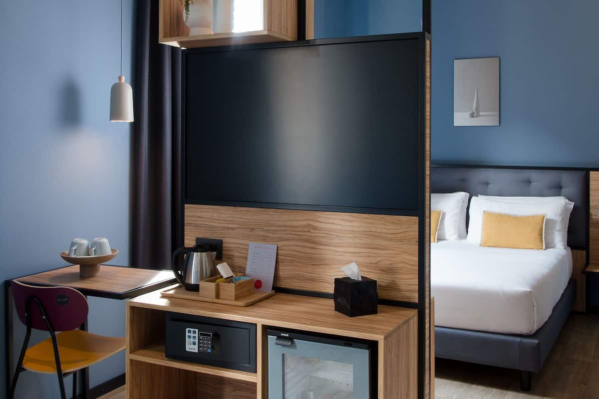 Een moderne hotelkamer met een bed, een bureau met een computer, een kleine kitchenette en een grote spiegel.