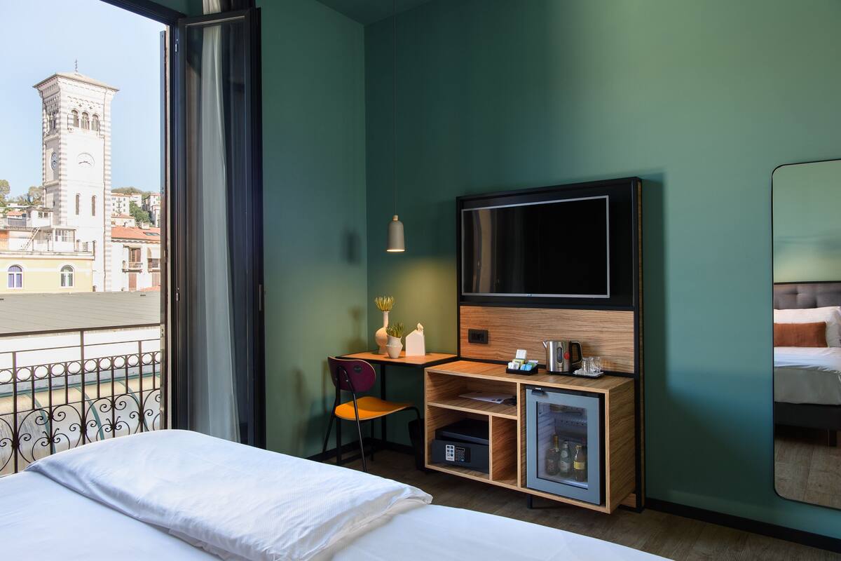 Hotelkamer met een bed, een bureau met een televisie, een kleine koelkast en een stoel. Een balkon met een hekwerk is zichtbaar.