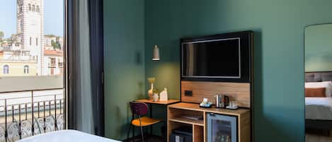 A hotel room with a bed, a desk with a TV, a small fridge, and a chair. A balcony with a railing is visible.