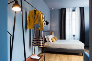 Una habitación de hotel moderna con una cama grande, una mesita de noche, un perchero con una chaqueta amarilla y una lámpara de pie.