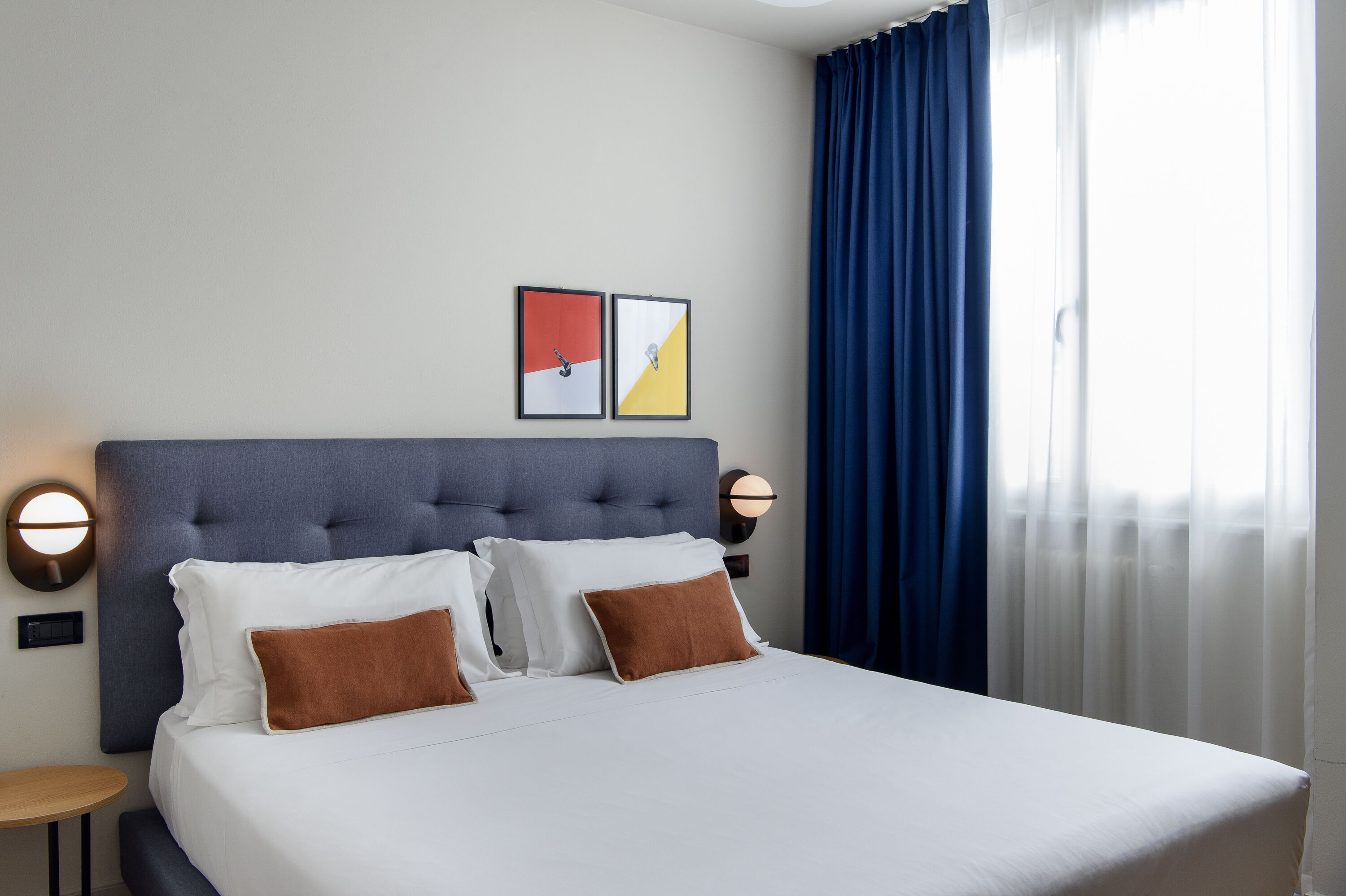A neatly made bed with white and brown pillows, a round wall clock, and two framed pictures on the wall.