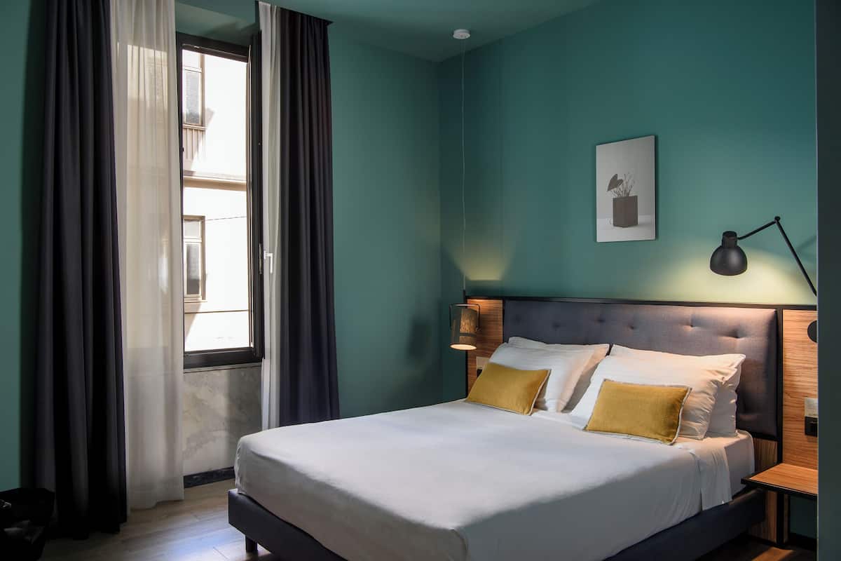 Een net opgemaakt bed met witte en gele kussens, een bedlamp en een ingelijst foto op de muur.