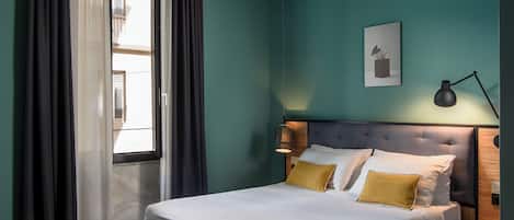 Una cama bien tendida con almohadas blancas y amarillas, una lámpara de noche y un cuadro en la pared.