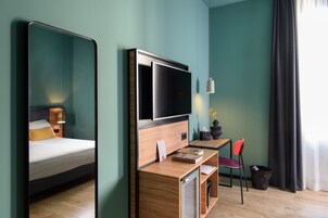 Habitación de hotel con cama, escritorio con silla, televisión y ventana con cortinas.