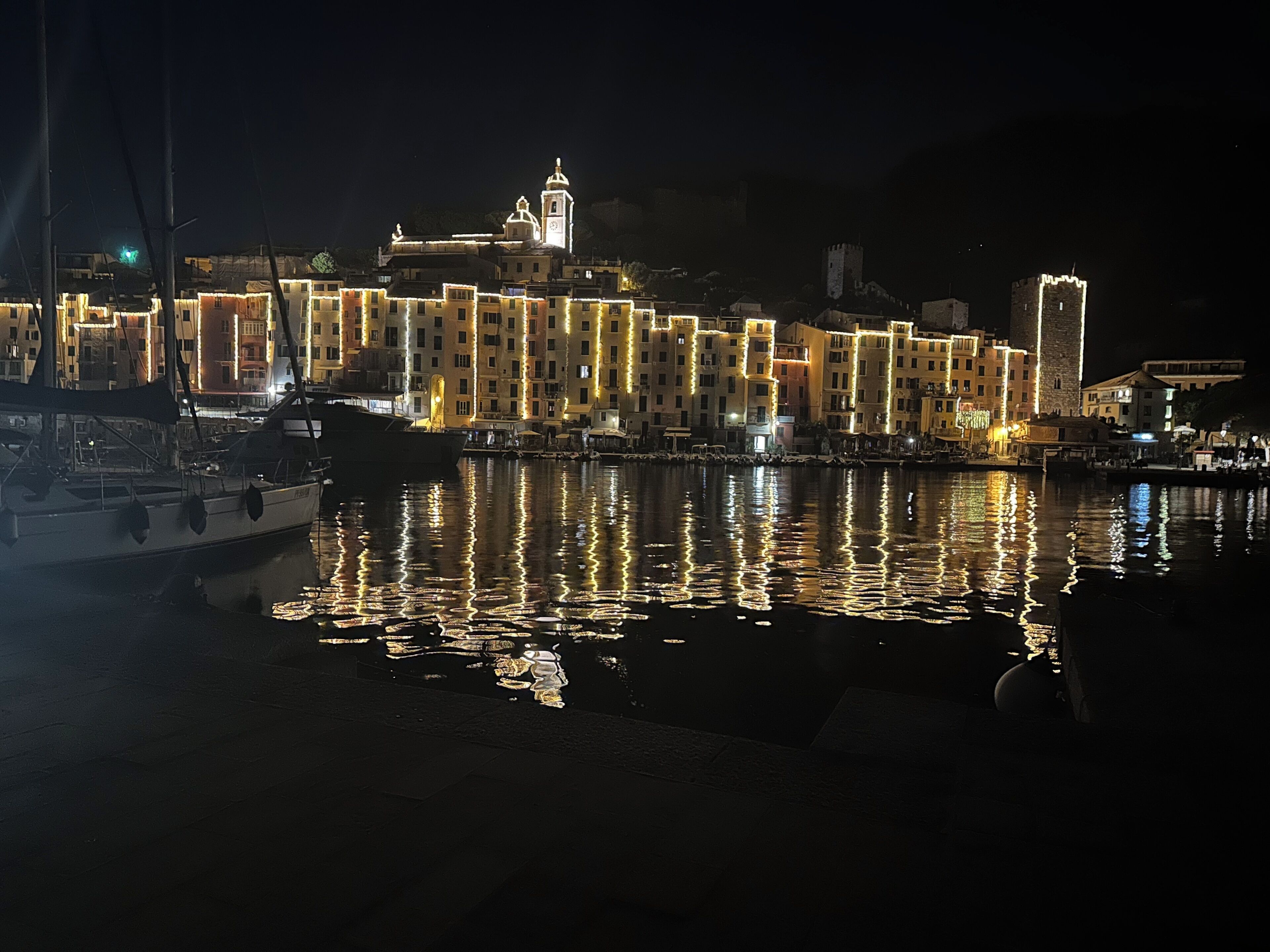 Paisagem urbana à beira-mar à noite, com edifícios iluminados e um barco atracado em primeiro plano.