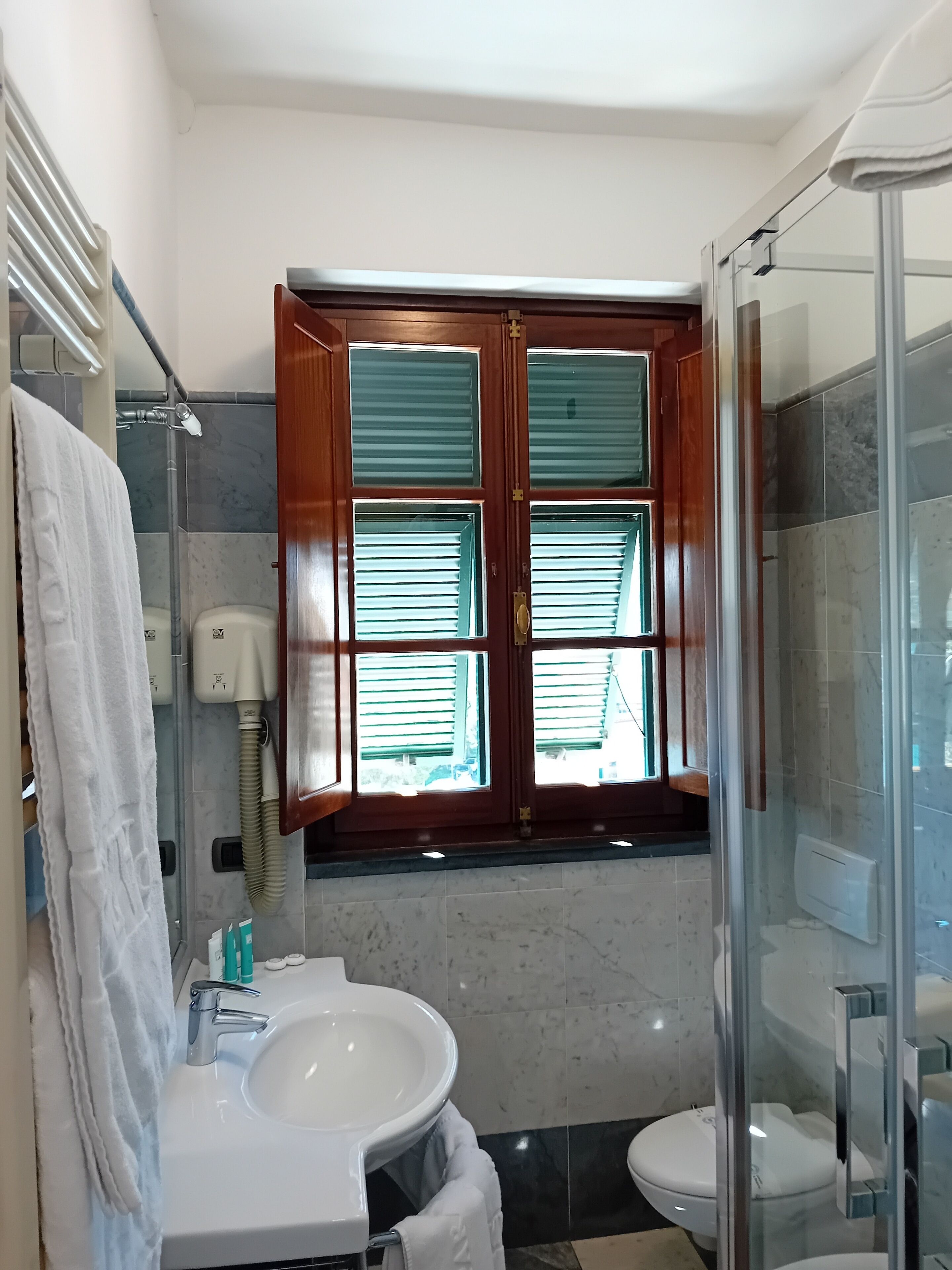 Baño con lavamanos blanco, inodoro y ducha con mampara de vidrio. Se aprecia una ventana con contraventanas de madera y una toalla colgada en la pared.