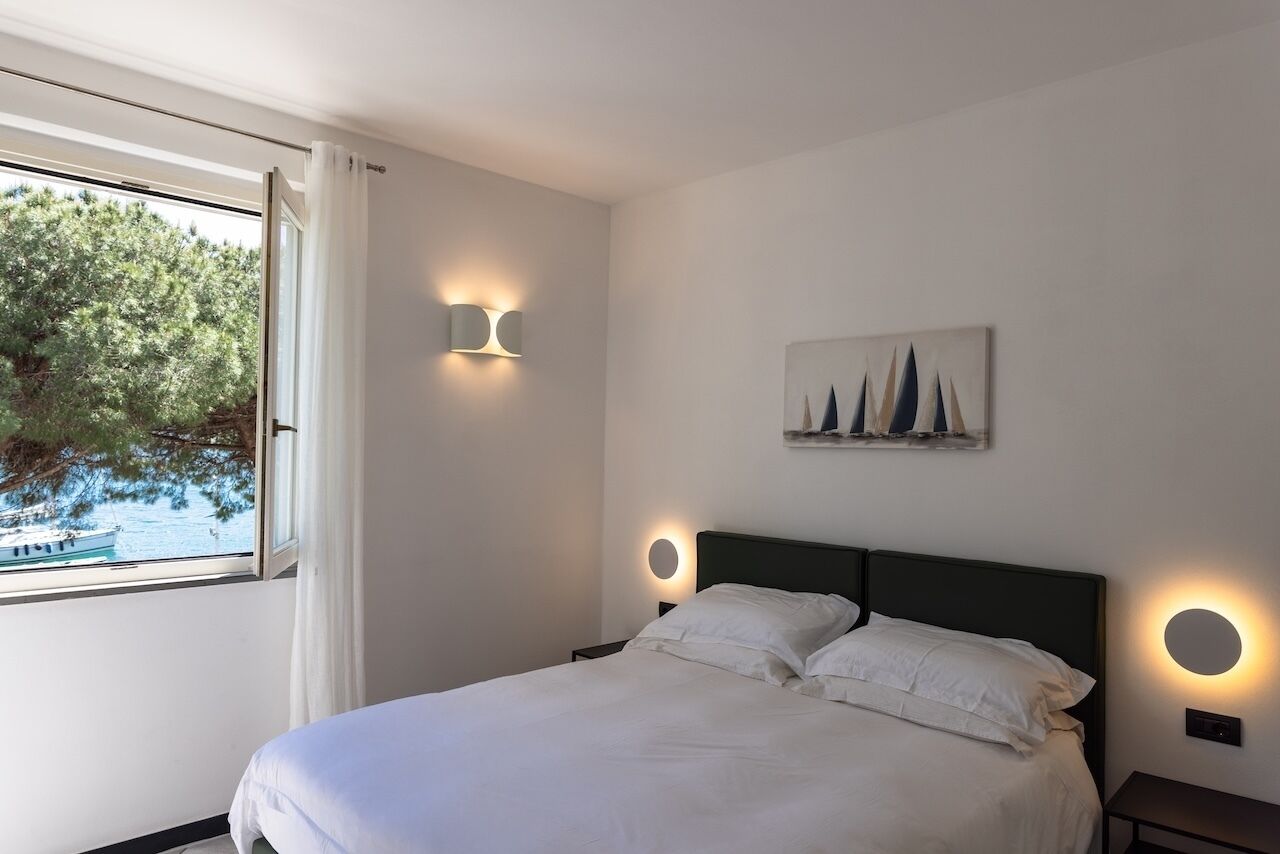 Une chambre à coucher comprenant un lit, deux tables de chevet, une lampe fixée au mur, un tableau et une fenêtre donnant sur des arbres et un bateau.