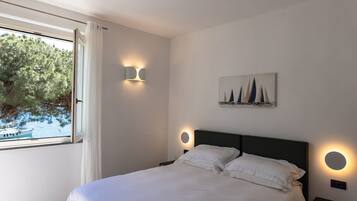 Une chambre à coucher comprenant un lit, deux tables de chevet, une lampe fixée au mur, un tableau et une fenêtre donnant sur des arbres et un bateau.
