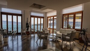 A spacious living room with large windows offering a view of the sea and mountains. There are wicker chairs, a white sofa, and a coffee table. - Hotel Paradiso (Portovenere)
