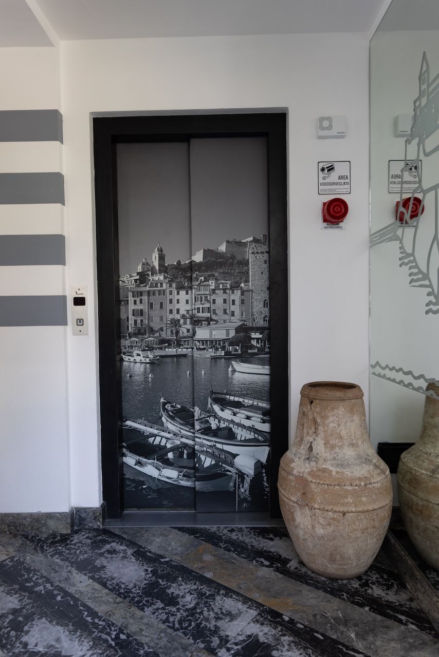 A door with a black and white photograph of a coastal town, a fire alarm, and a large vase on the floor.
