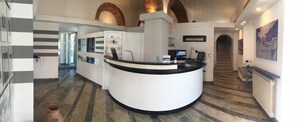 Un área de recepción con un mostrador blanco curvo, una computadora y un escritorio con una silla. Hay una escalera que conduce a un nivel superior y un cuadro grande en la pared.