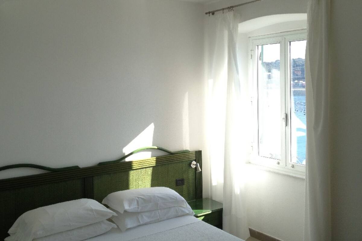 A bedroom with a bed, a nightstand, and a window with a view of the sea.