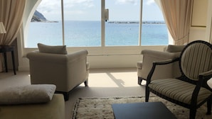 A room with a large arched window overlooking the sea, featuring two wicker chairs and a small table.