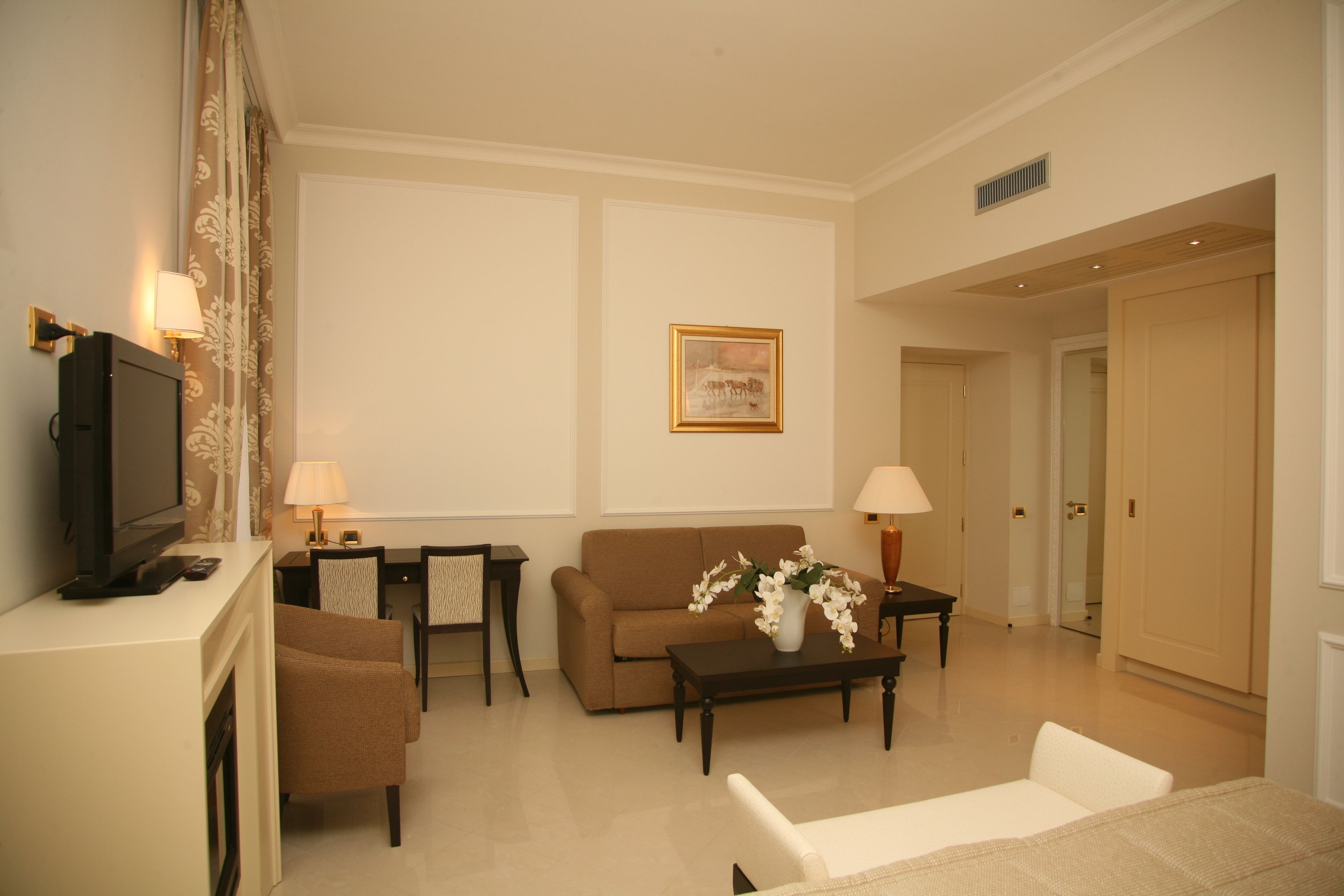 A hotel room with a TV, sofa, armchair, coffee table, and a painting on the wall.