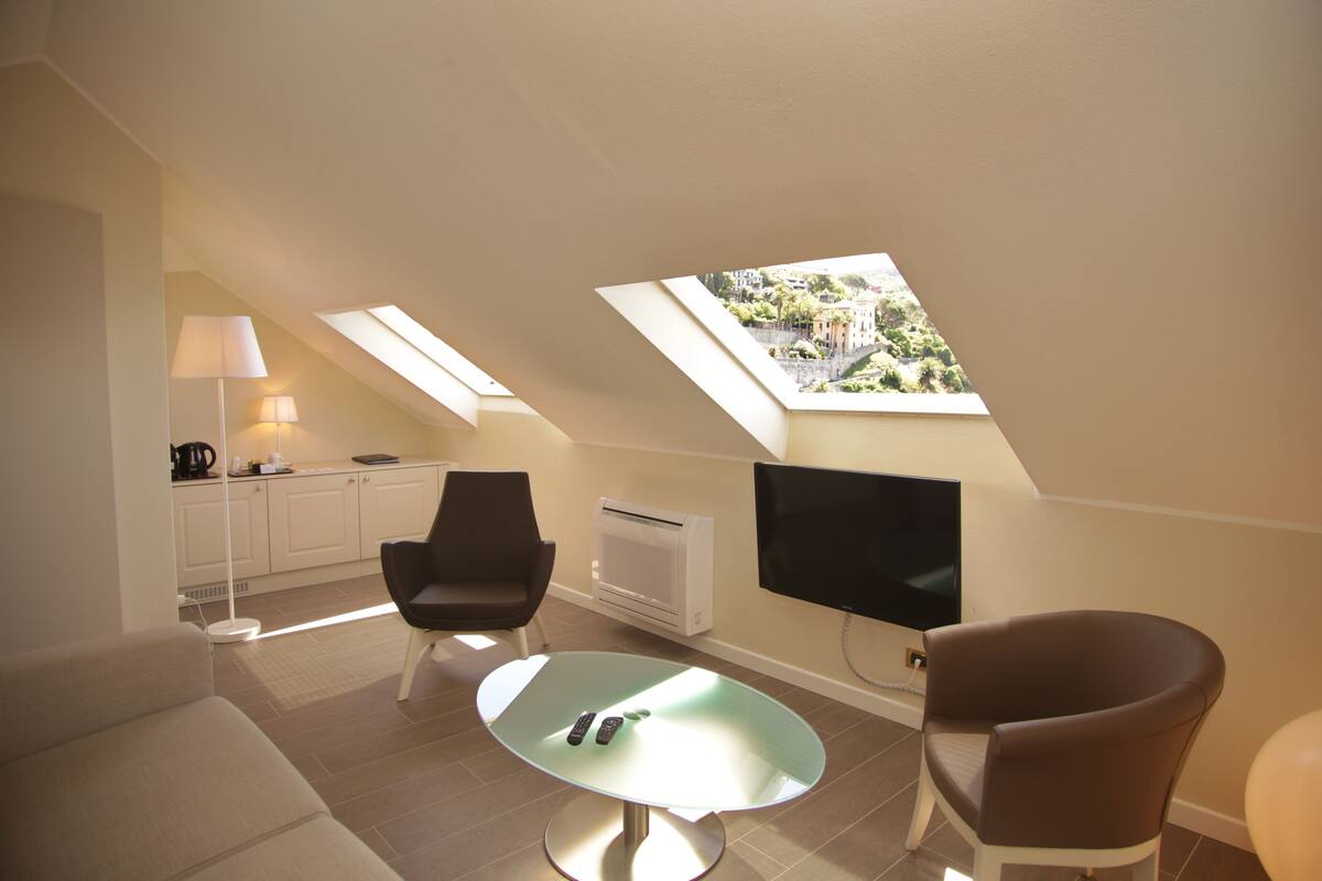 Een moderne woonkamer met een glazen tafel, twee fauteuils, een bank, een flatscreen televisie en een keukenhoek met een lamp.