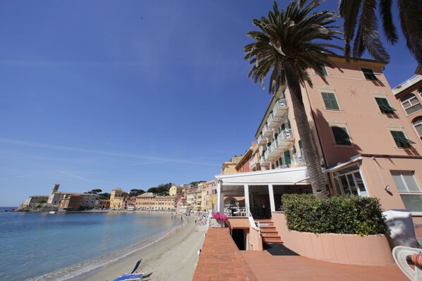 A beachfront hotel with a terrace, palm trees, and a clear blue sky.