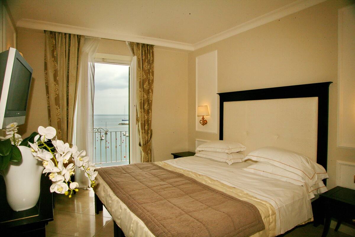 Een hotelkamer met een bed, een nachtkastje, een televisie, een vaas met bloemen en uitzicht op zee door een raam.