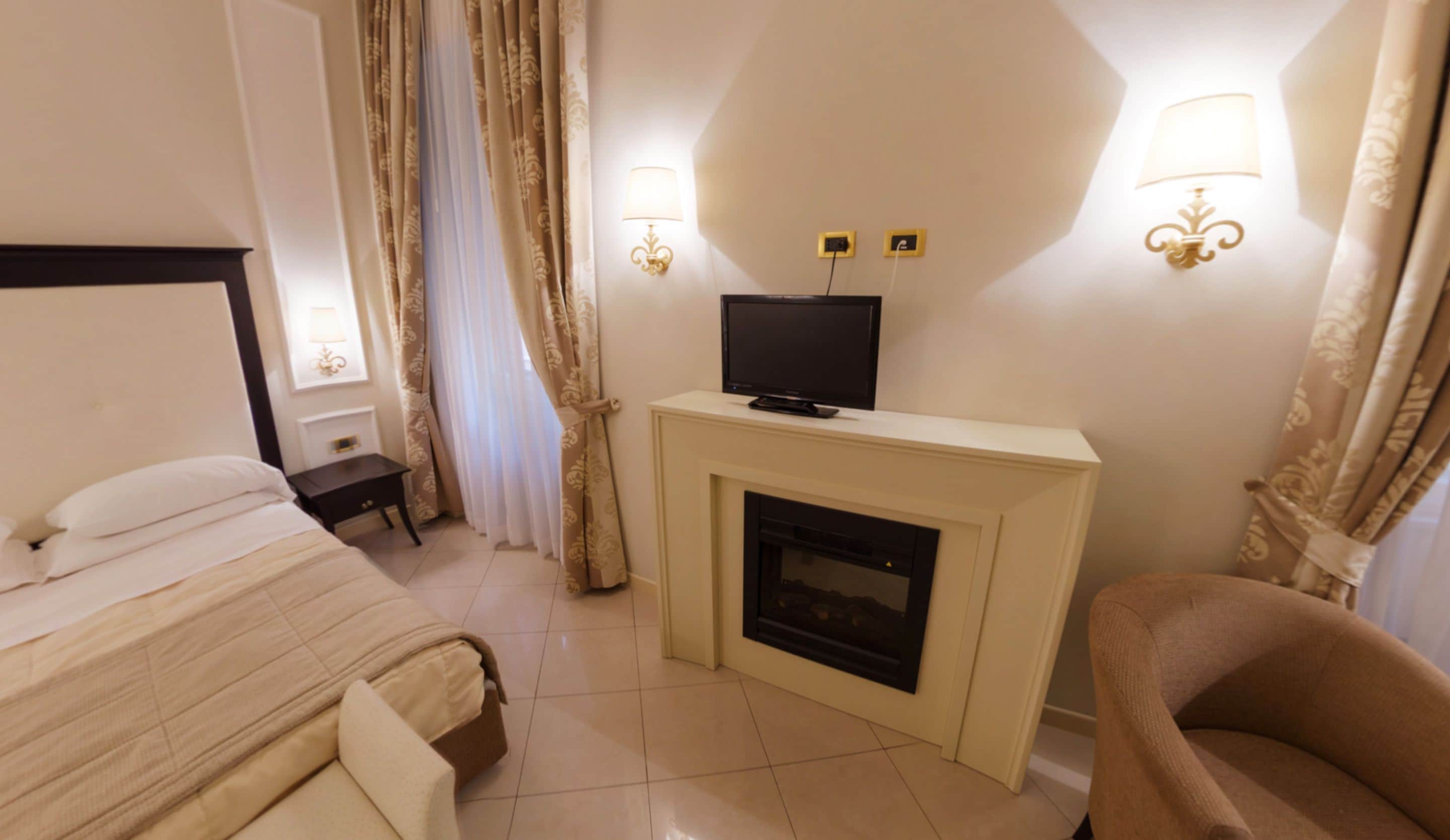 A hotel room with a bed, a television on a stand, a fireplace, a nightstand, and two wall-mounted lamps.