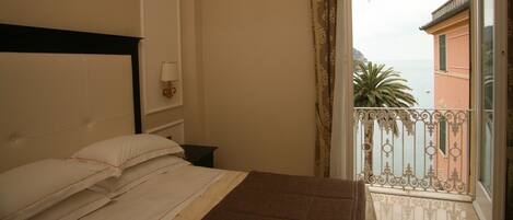 A bedroom with a bed, a nightstand, a wall-mounted lamp, and a view of the sea through a balcony with a decorative railing.