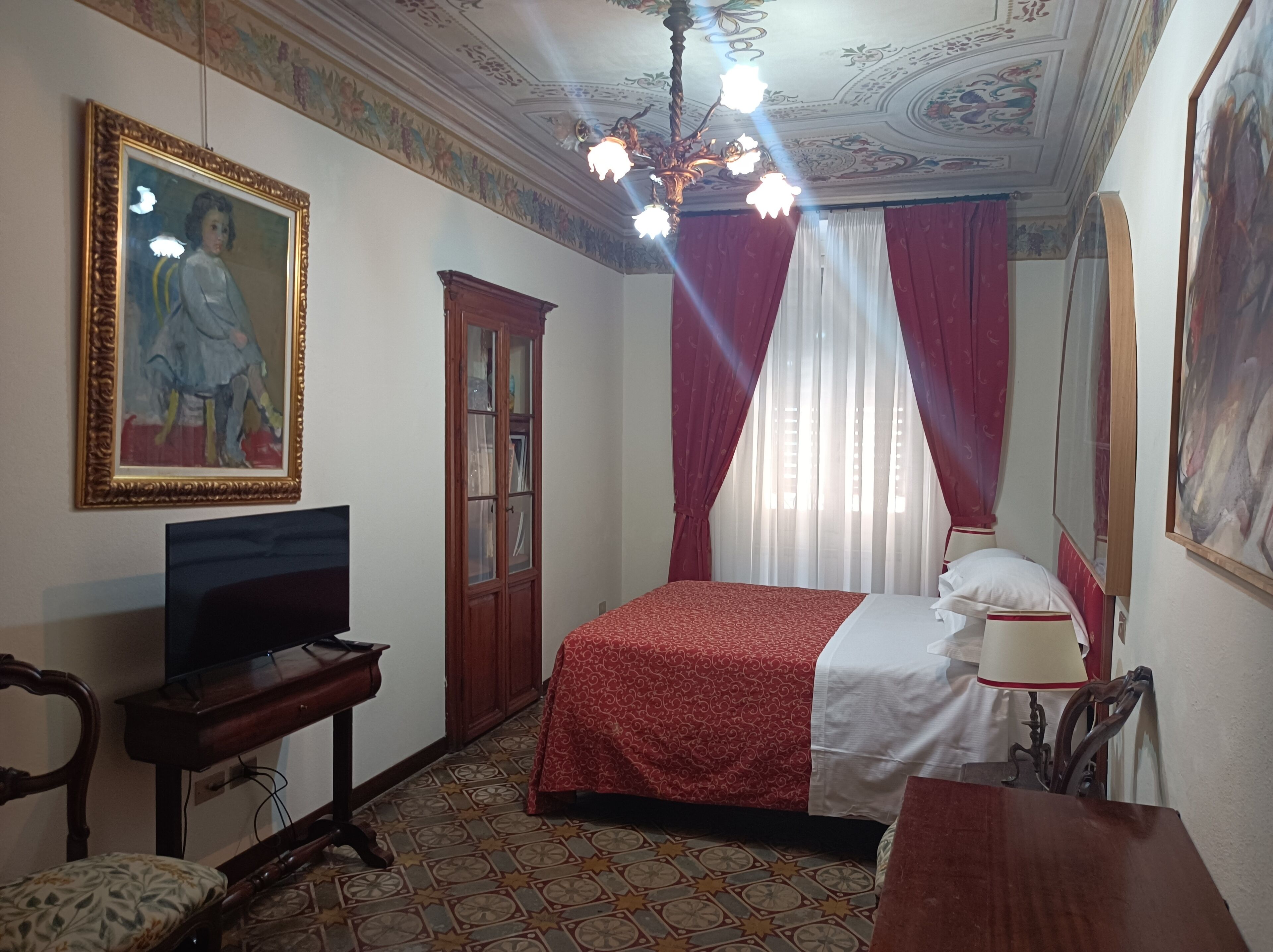Ein Hotelzimmer mit einem Bett, einem Fernseher, einem Holzschrank, einem Sessel, einem Schreibtisch und einer Decke mit ornamentalem Dekor.
