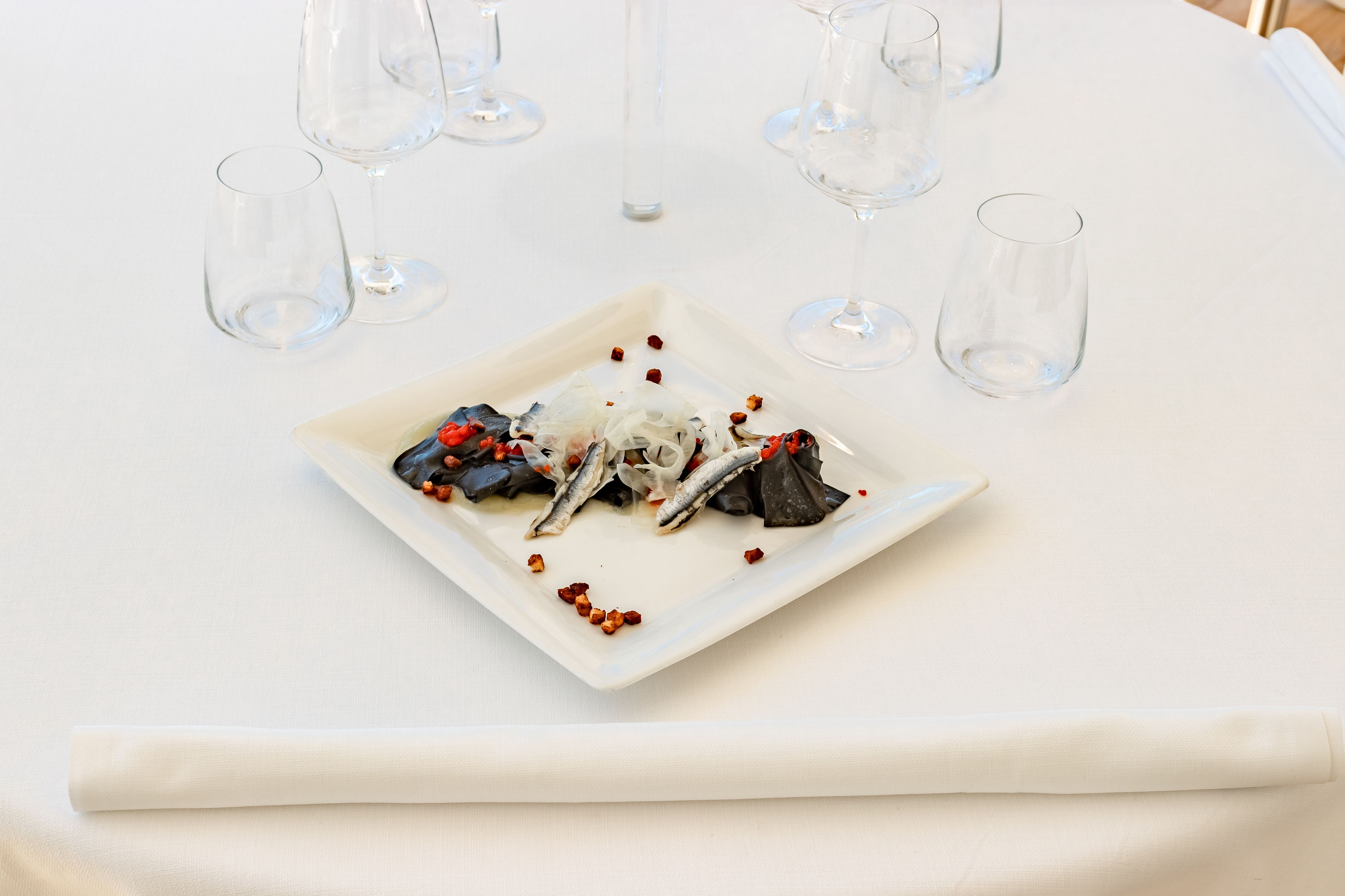 A square white plate with a seafood dish, surrounded by empty wine glasses on a white tablecloth.
