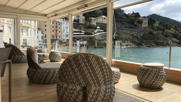 A modern lounge area with wicker furniture, large windows offering a view of a coastal town and water.