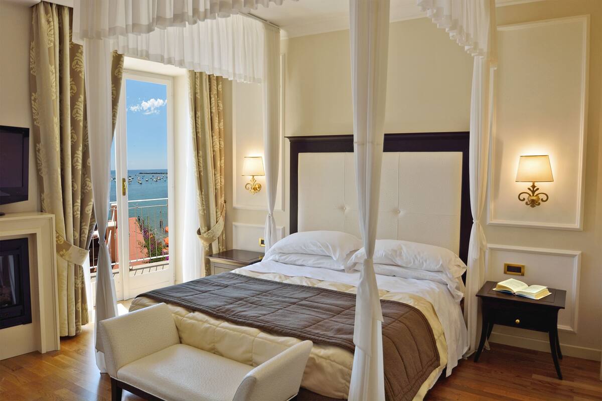 Een slaapkamer met een hemelbed, een zithoek met een stoel, een nachtkastje met een lamp en uitzicht op zee door een raam.
