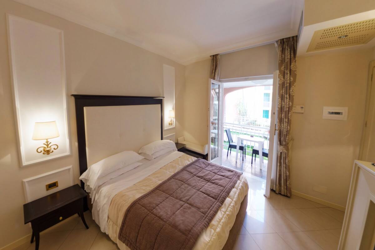 Hotelkamer met een bed, nachtkastje, lamp, raam met gordijnen en uitzicht op het balkon.
